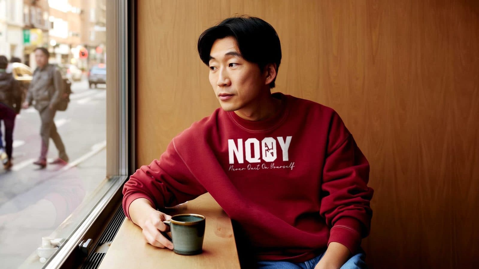 A man wearing an NQOY cardinal crewneck at a coffee shop window